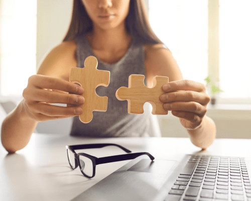 Person holding two wooden puzzle pieces near a laptop, symbolizing system integration or connecting tools in a digital environment.