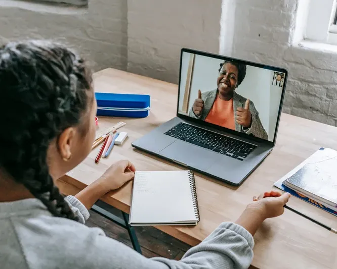 A student sits at a desk with a notebook and laptop, smiling at a teacher giving a thumbs-up during an online tutoring session.