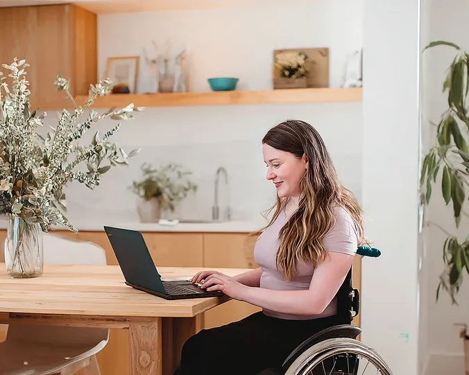 A woman in a wheelchair sitting at a wooden table using a laptop to take an online exam in a bright home environment.