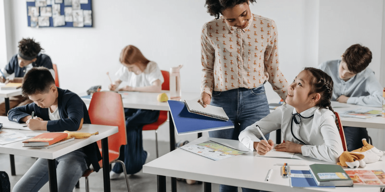 Teacher engaging with a smiling student in a primary classroom while other students are working at their desks on school assignments.