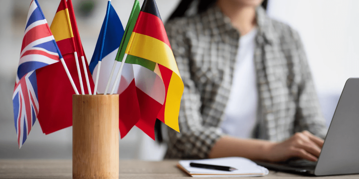 A desk with small flags of various countries including the UK, Spain, France, and Germany in a wooden holder, with a smiling woman typing on a laptop in the background, symbolizing language learning and international communication.