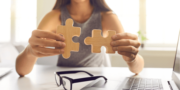 Person holding two wooden puzzle pieces near a laptop, symbolizing system integration or connecting tools in a digital environment.