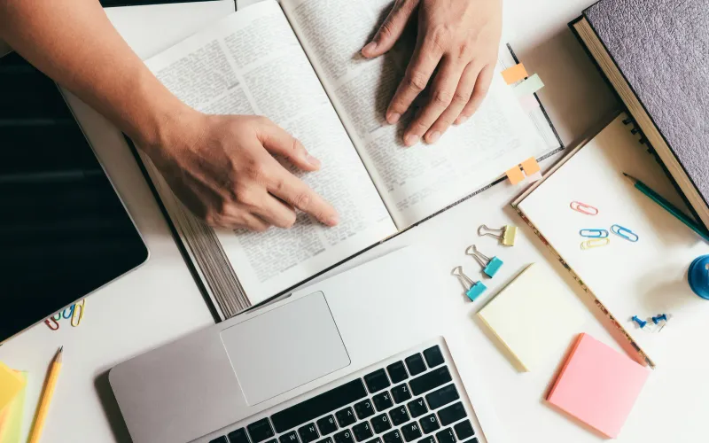 Student studying with open books, a laptop, and stationery supplies on a desk, representing preparation and academic assessment.