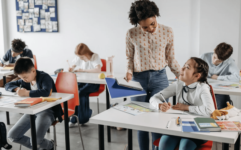 Teacher engaging with a smiling student in a primary classroom while other students are working at their desks on school assignments.