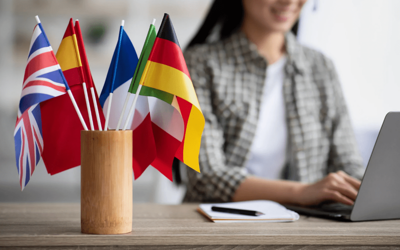 A desk with small flags of various countries including the UK, Spain, France, and Germany in a wooden holder, with a smiling woman typing on a laptop in the background, symbolizing language learning and international communication.