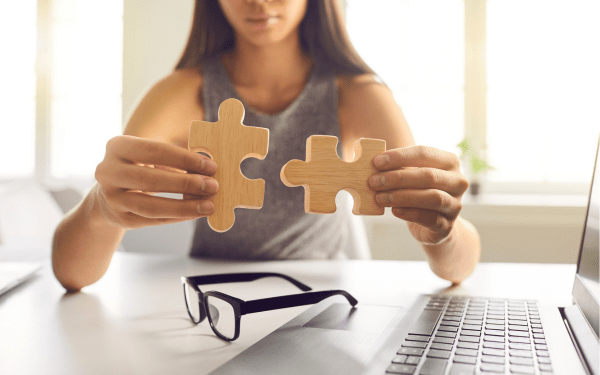 Person holding two wooden puzzle pieces near a laptop, symbolizing system integration or connecting tools in a digital environment.
