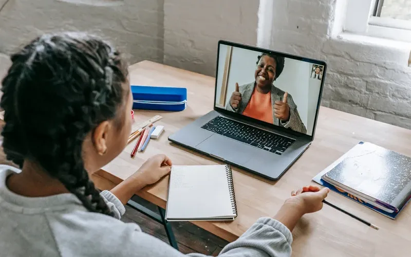 A student sits at a desk with a notebook and laptop, smiling at a teacher giving a thumbs-up during an online tutoring session.