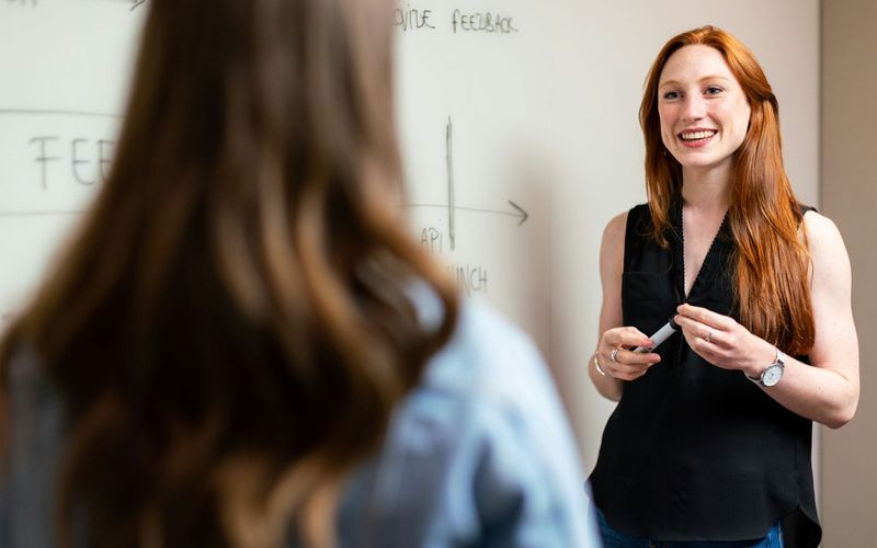 A tutor enthusiastically explaining concepts to a student in a modern educational environment. The tutor stands in front of a whiteboard, smiling and engaging in a discussion.