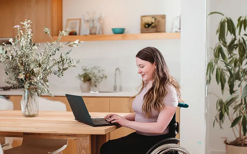 A woman in a wheelchair sitting at a wooden table using a laptop to take an online exam in a bright home environment.