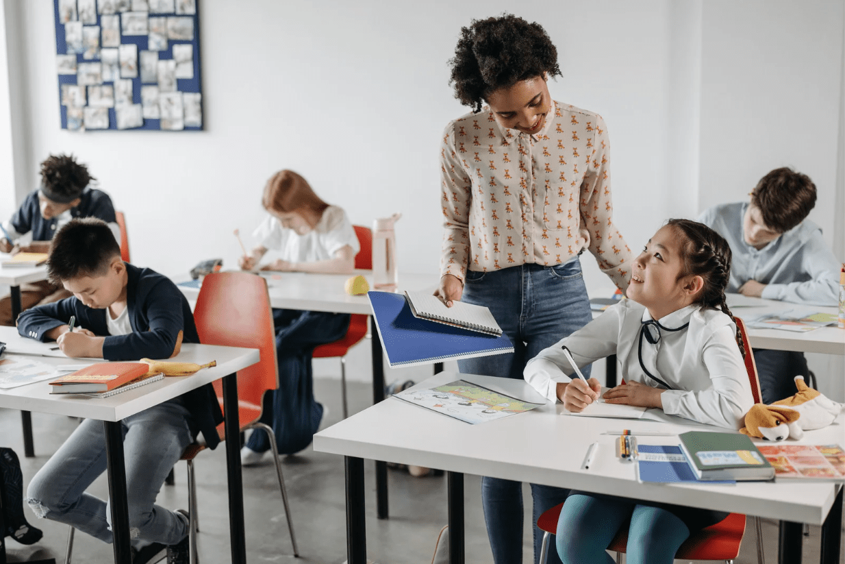 Teacher engaging with a smiling student in a primary classroom while other students are working at their desks on school assignments.