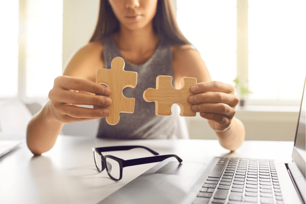 Person holding two wooden puzzle pieces near a laptop, symbolizing system integration or connecting tools in a digital environment.