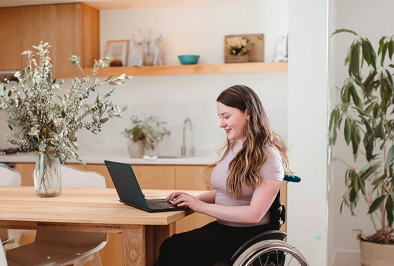 A woman in a wheelchair sitting at a wooden table using a laptop to take an online exam in a bright home environment.